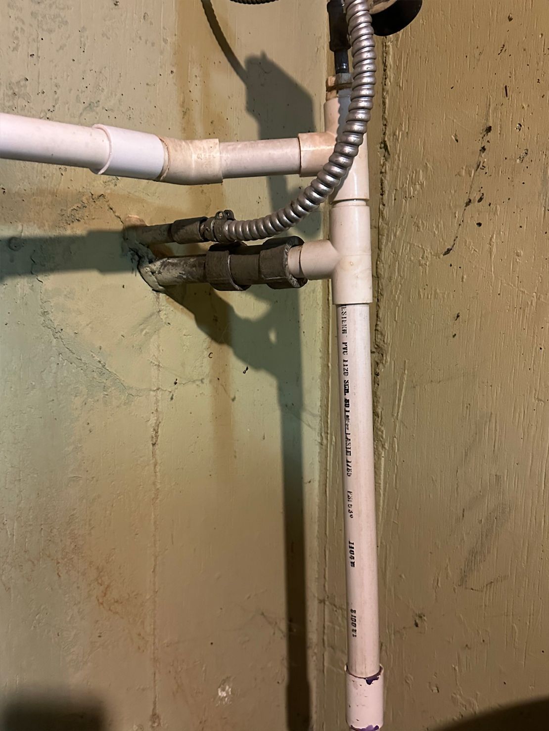 White PVC pipes and valves on a stained concrete wall; electrical conduit runs alongside the pipes.