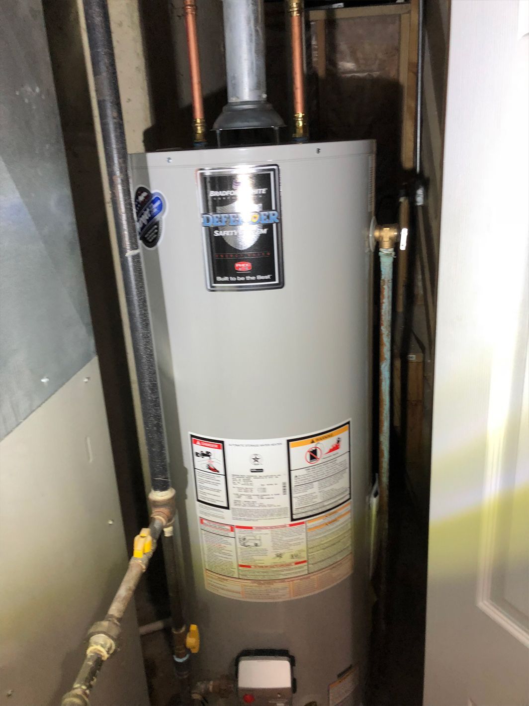 Gray electric water heater in a utility room, with pipes and labels visible.