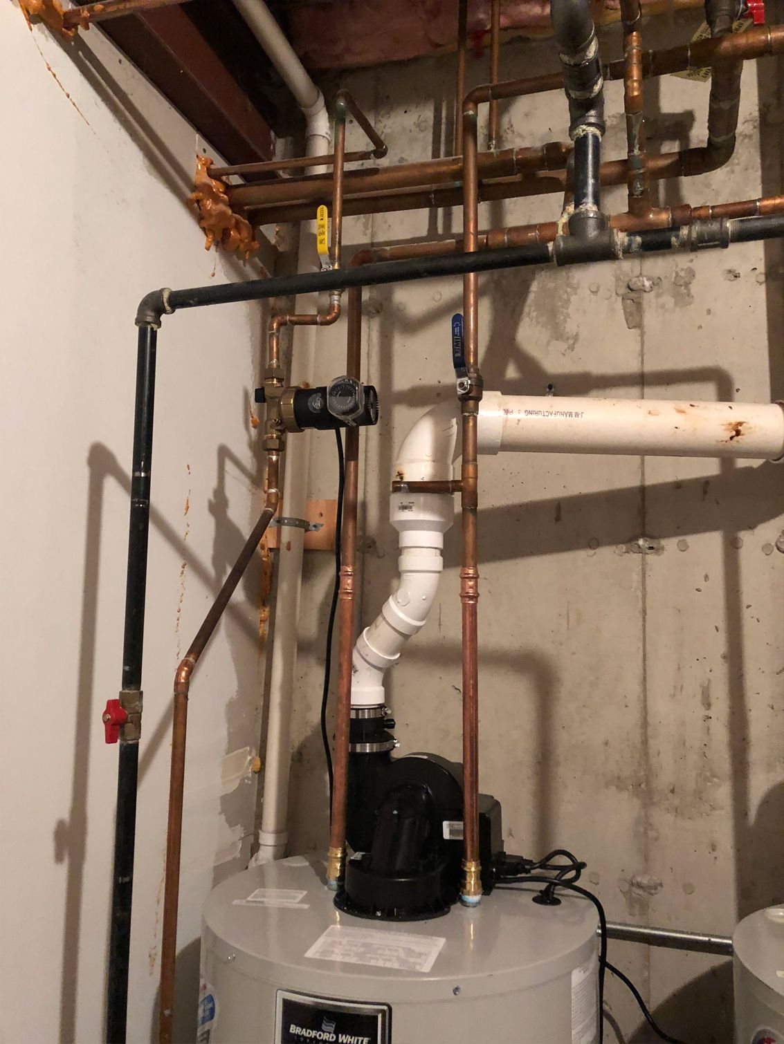 Plumbing pipes above a hot water heater in a basement, including copper, black, and white pipes.