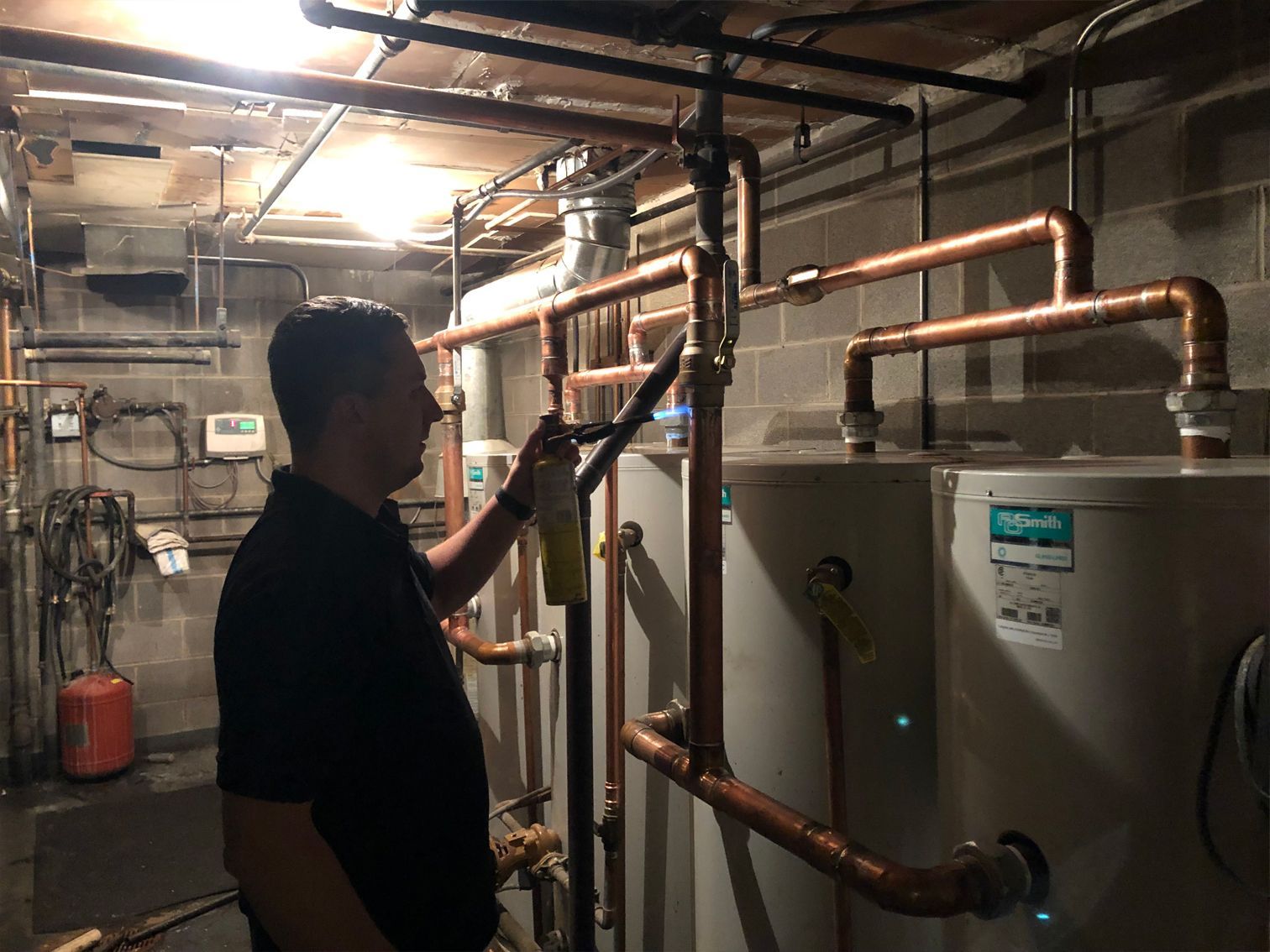 Plumber using a torch on copper pipes near water heaters in a basement.