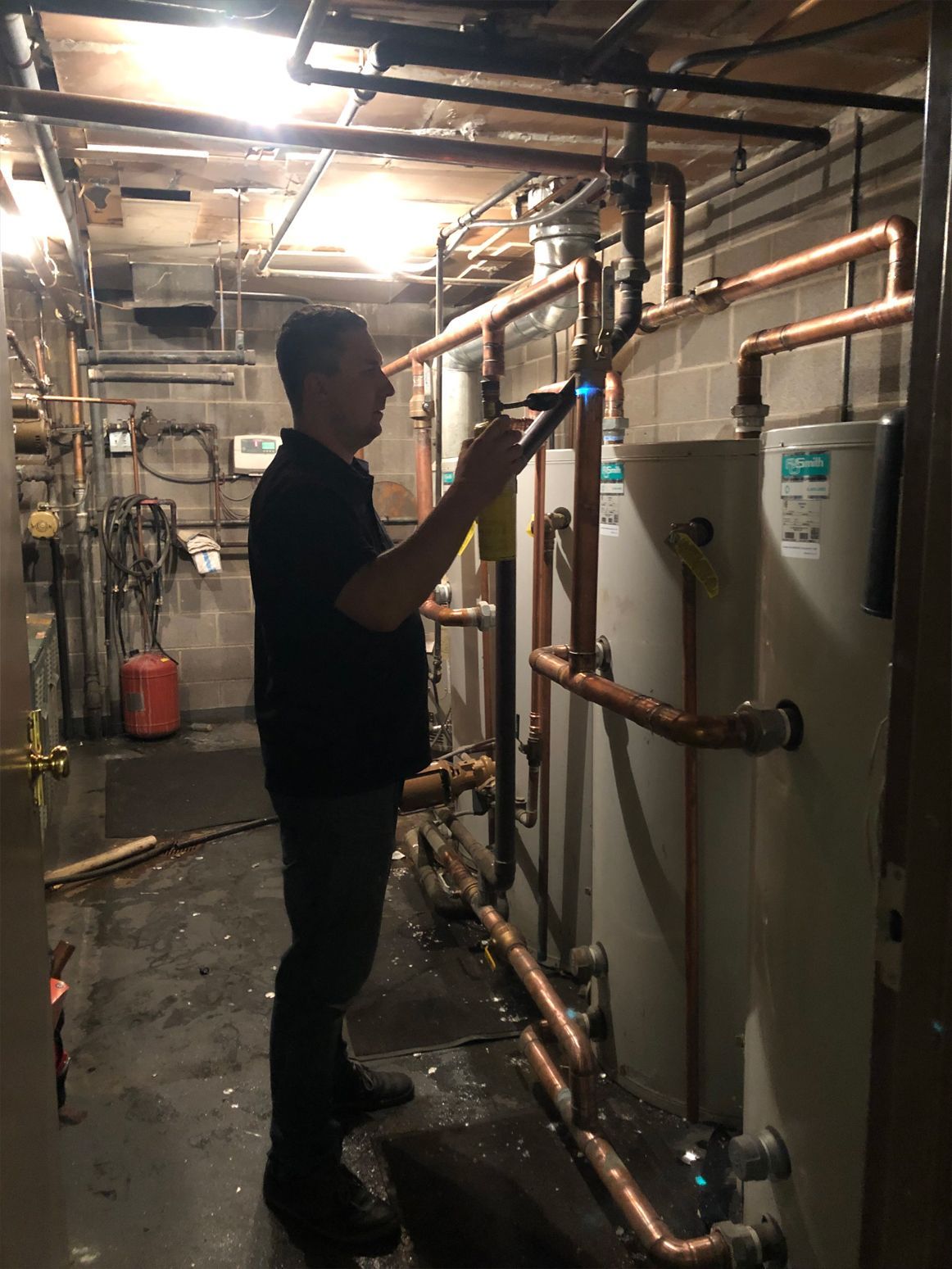 Man in a black shirt uses a torch on copper pipes near water heaters in a basement.