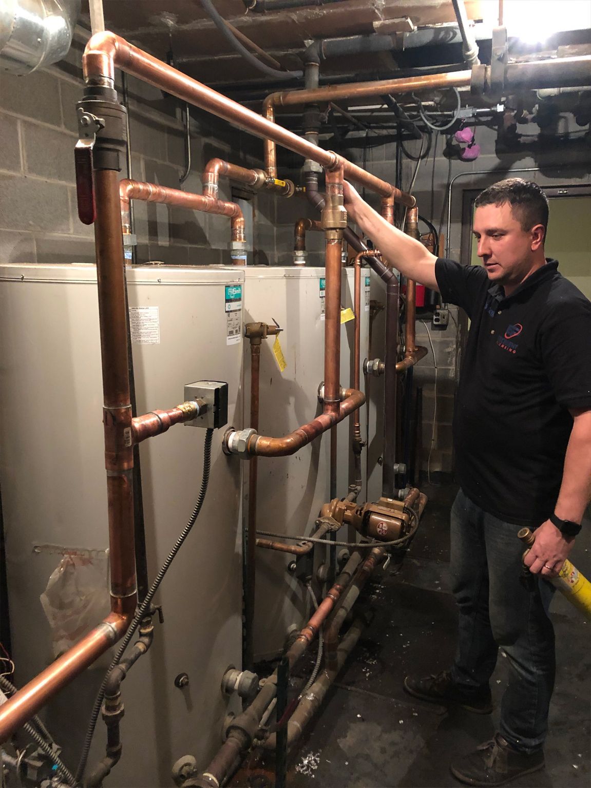 Man in a black shirt examining copper pipes connected to a water heater in a basement.
