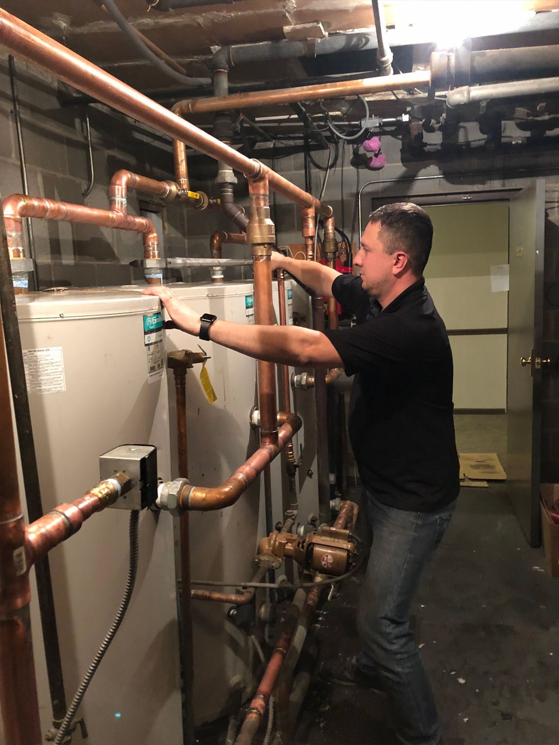 Man inspecting pipes on a heating system in a basement; copper pipes, dark shirt, jeans.