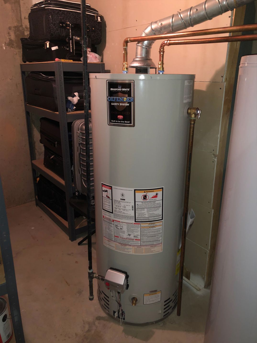 A water heater in a basement. It's a tall, gray cylindrical tank with copper pipes connected.