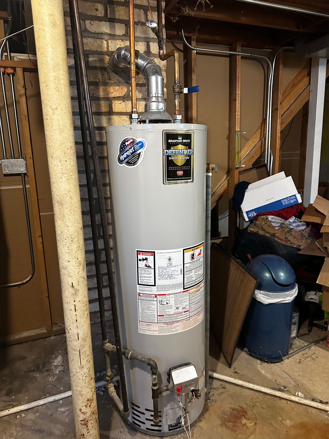 A tall gray water heater in a basement setting, with pipes and a chimney.