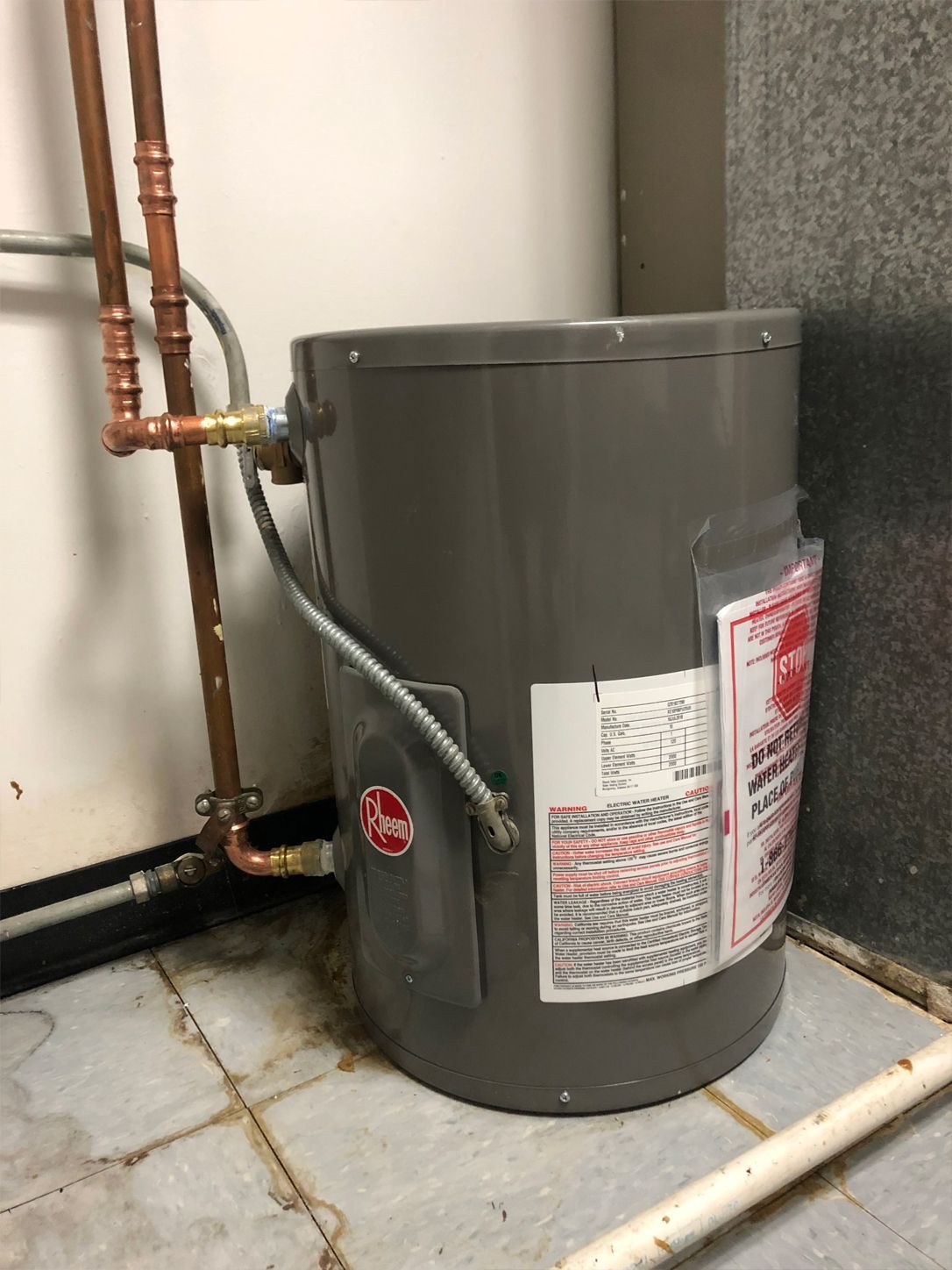 A Rheem electric water heater against a white wall and a tiled floor. Copper pipes are connected.