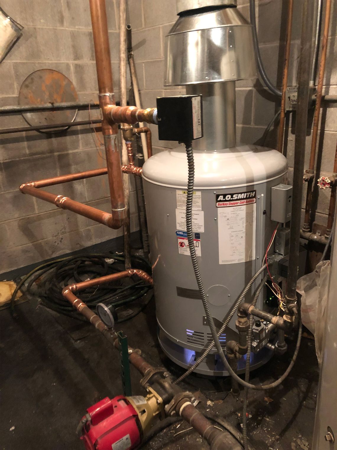 Water heater with copper pipes in a basement setting. A red pump sits on the floor.