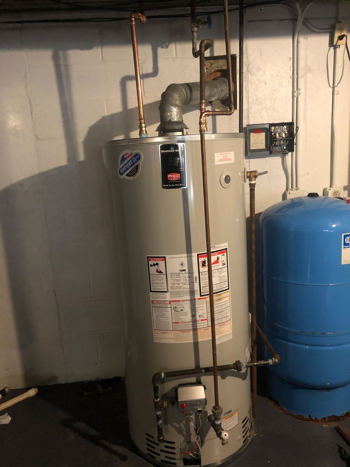 Gray water heater with copper pipes in a basement next to a blue tank.