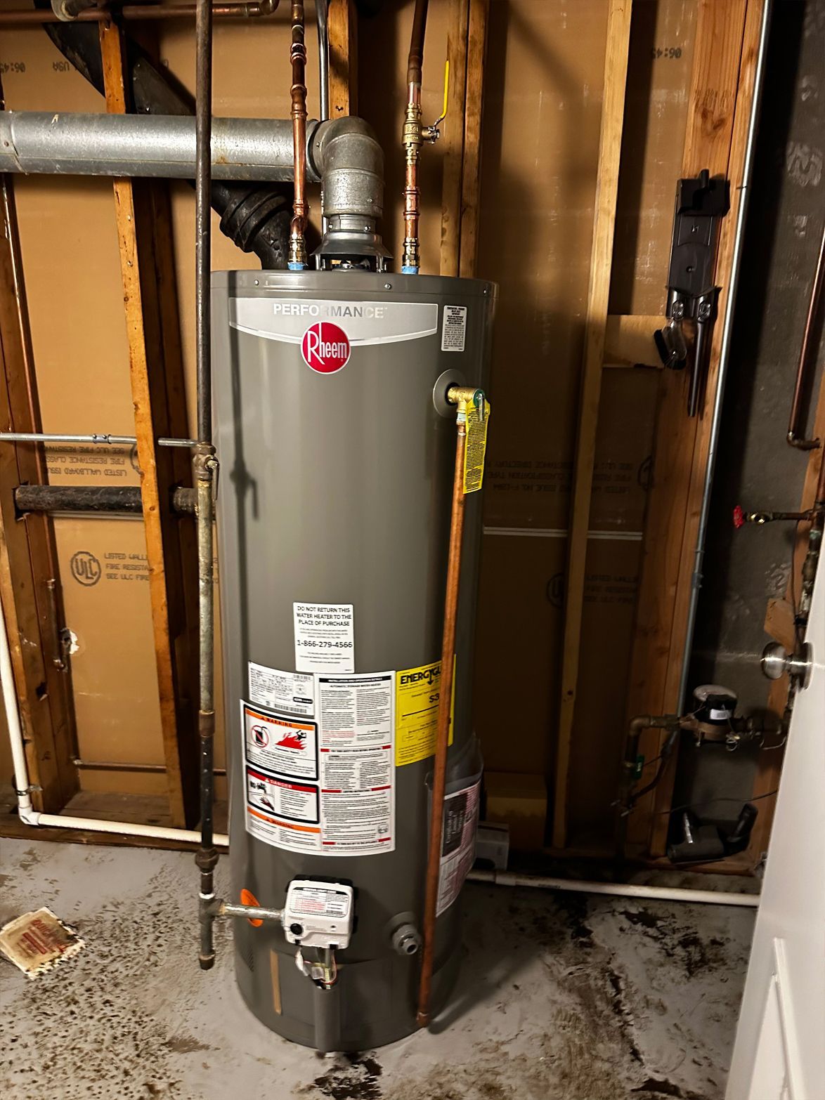 A Rheem brand water heater stands in a basement against wooden walls and concrete floor.