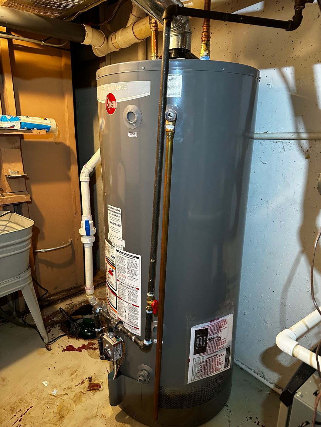 Tall, gray Rheem water heater in a basement. Copper pipes and water lines are attached.