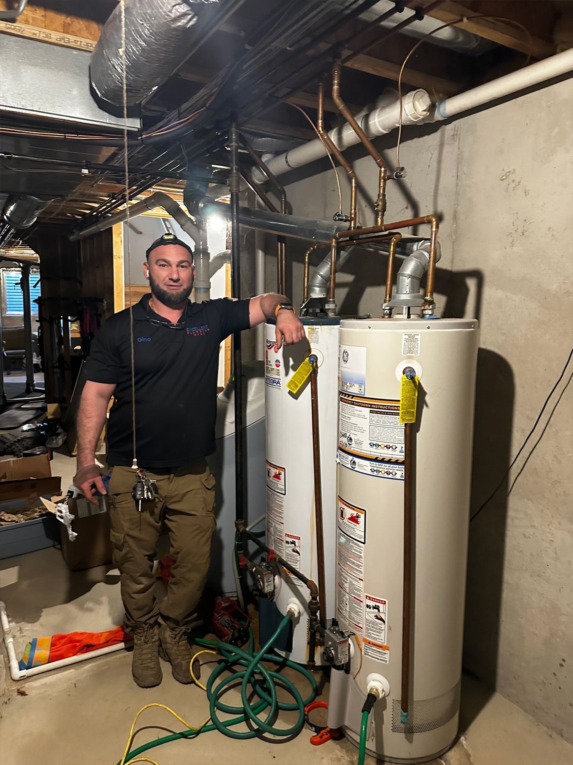 Man stands by two water heaters in a basement. Copper pipes visible. Green and yellow cords on floor.