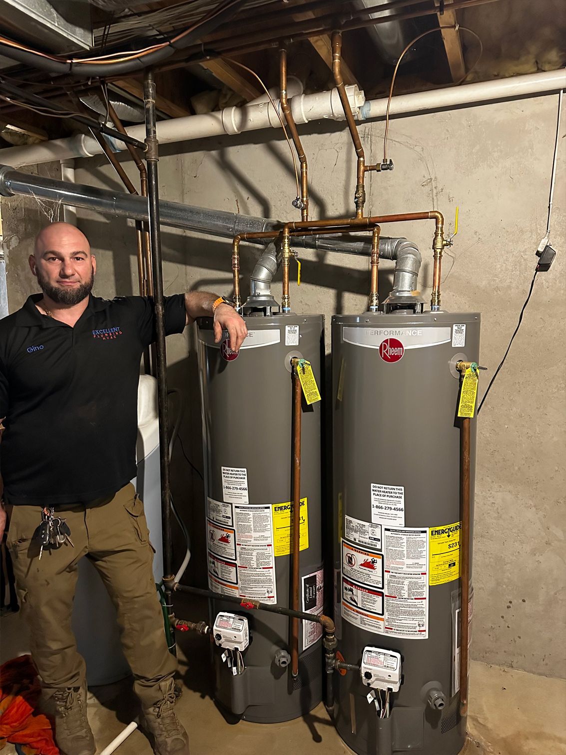 Man standing next to two gray water heaters in a basement setting, copper pipes overhead.