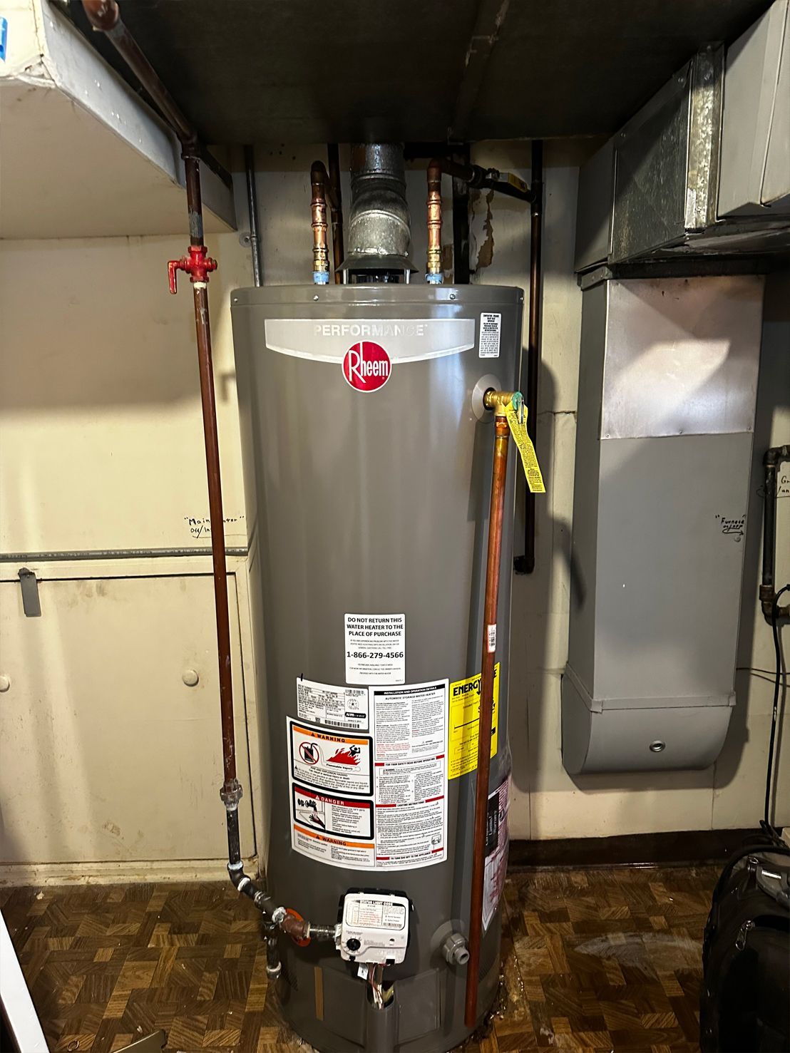 A Rheem water heater in a basement. It's gray and has copper pipes and a yellow tag.