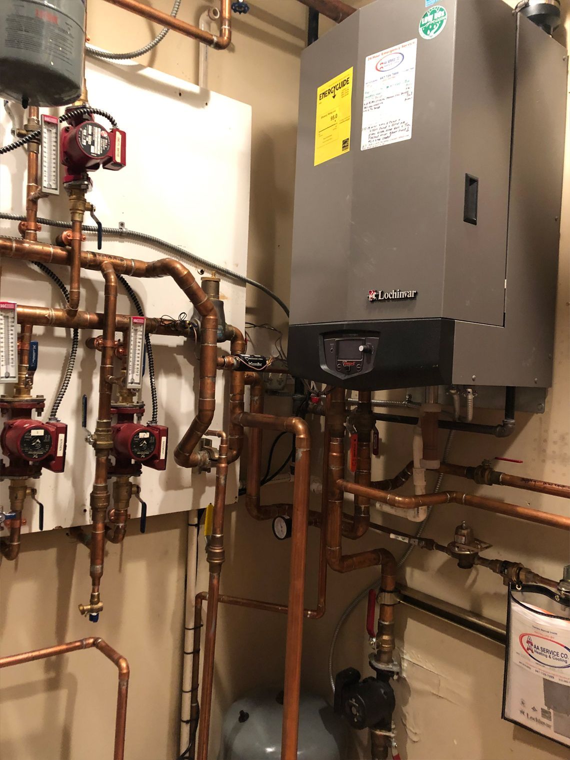Copper pipes and a gray boiler with red pumps against a tan wall in a utility room.