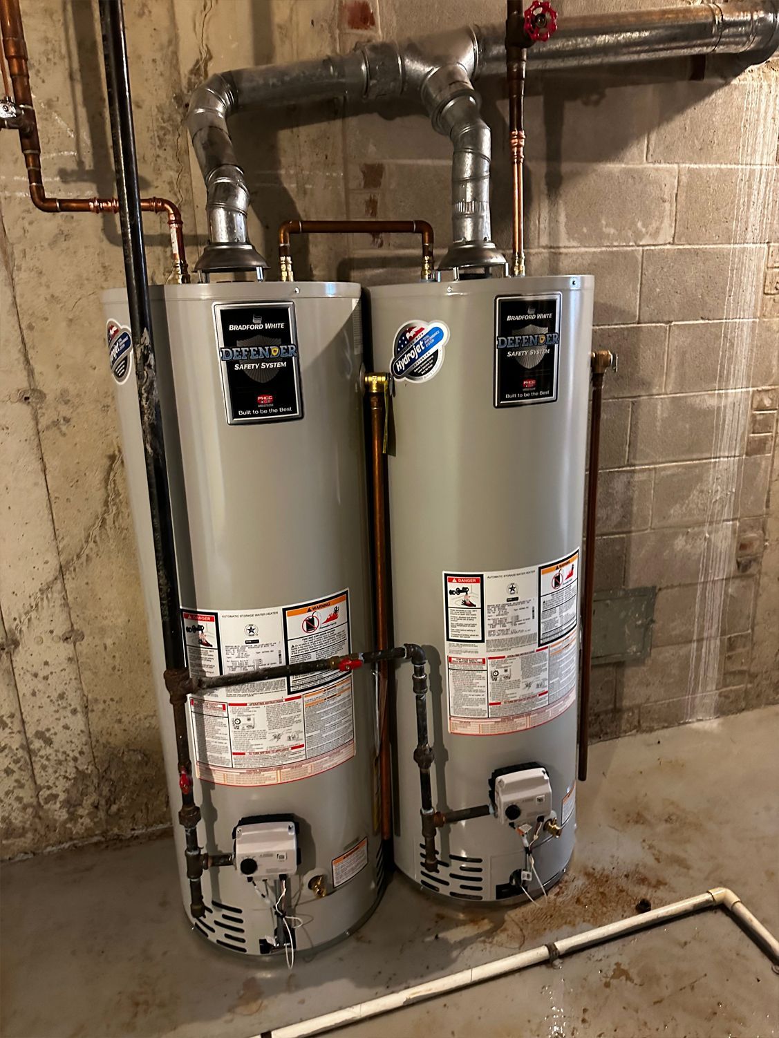 Two tall gray water heaters side-by-side in a basement, connected by pipes against a concrete block wall.