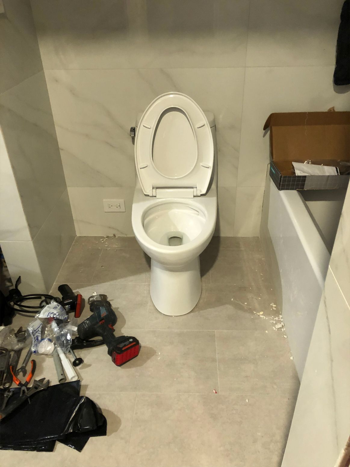White toilet with the seat up, tools on the floor. Bathroom setting with light gray tiles and a partially visible wall.