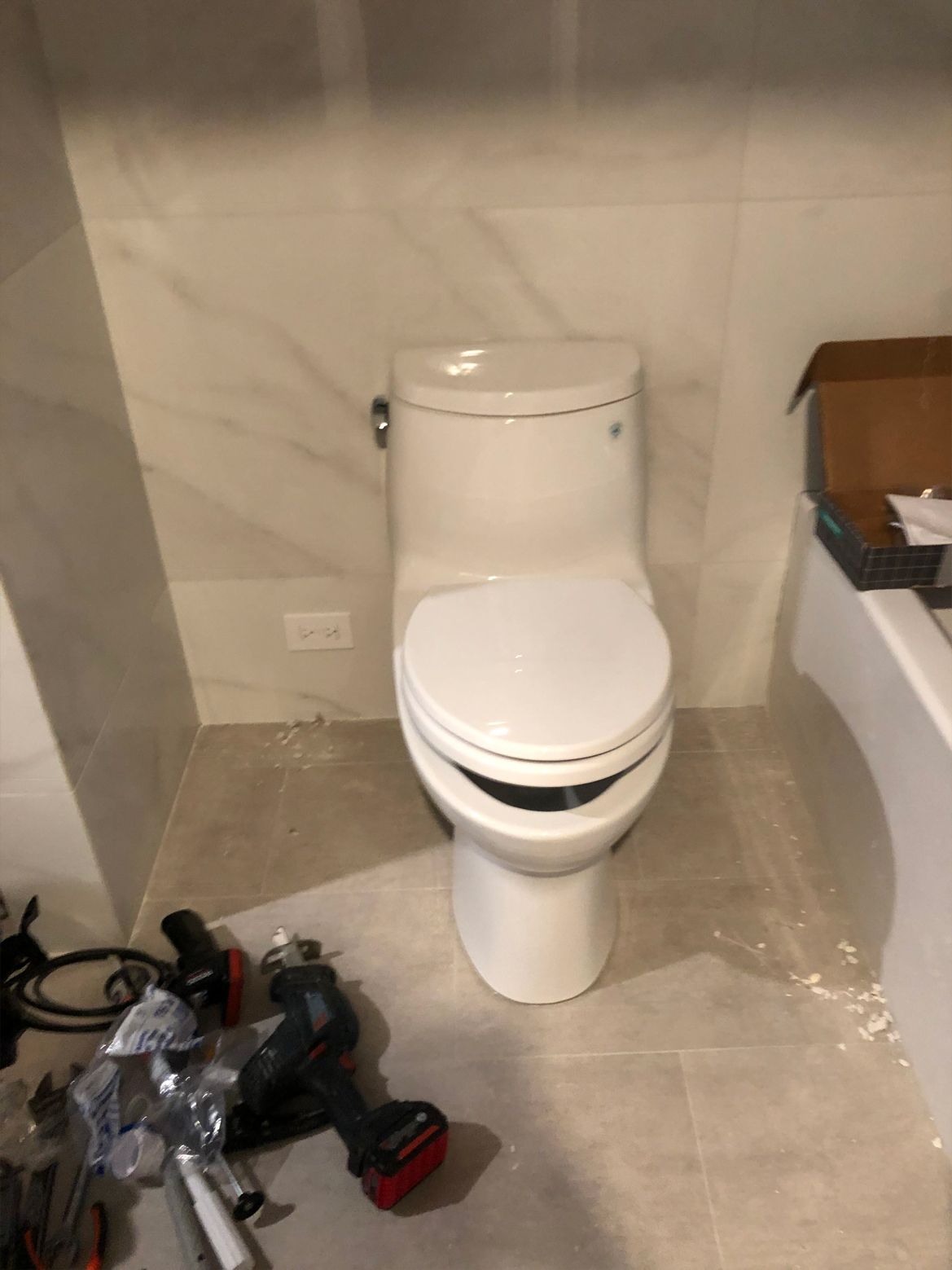 White toilet installed against a tiled wall in a bathroom, construction tools on the floor.