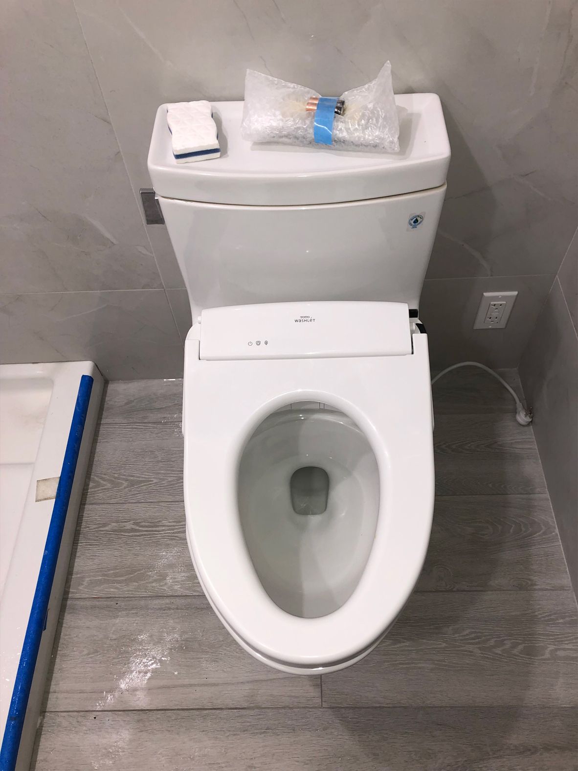 White toilet with a bidet seat in a bathroom. Grey walls and flooring. Small items on the tank.