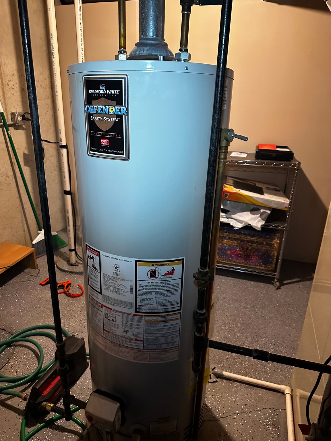 A cylindrical water heater with pipes and safety labels. It is in a basement setting.