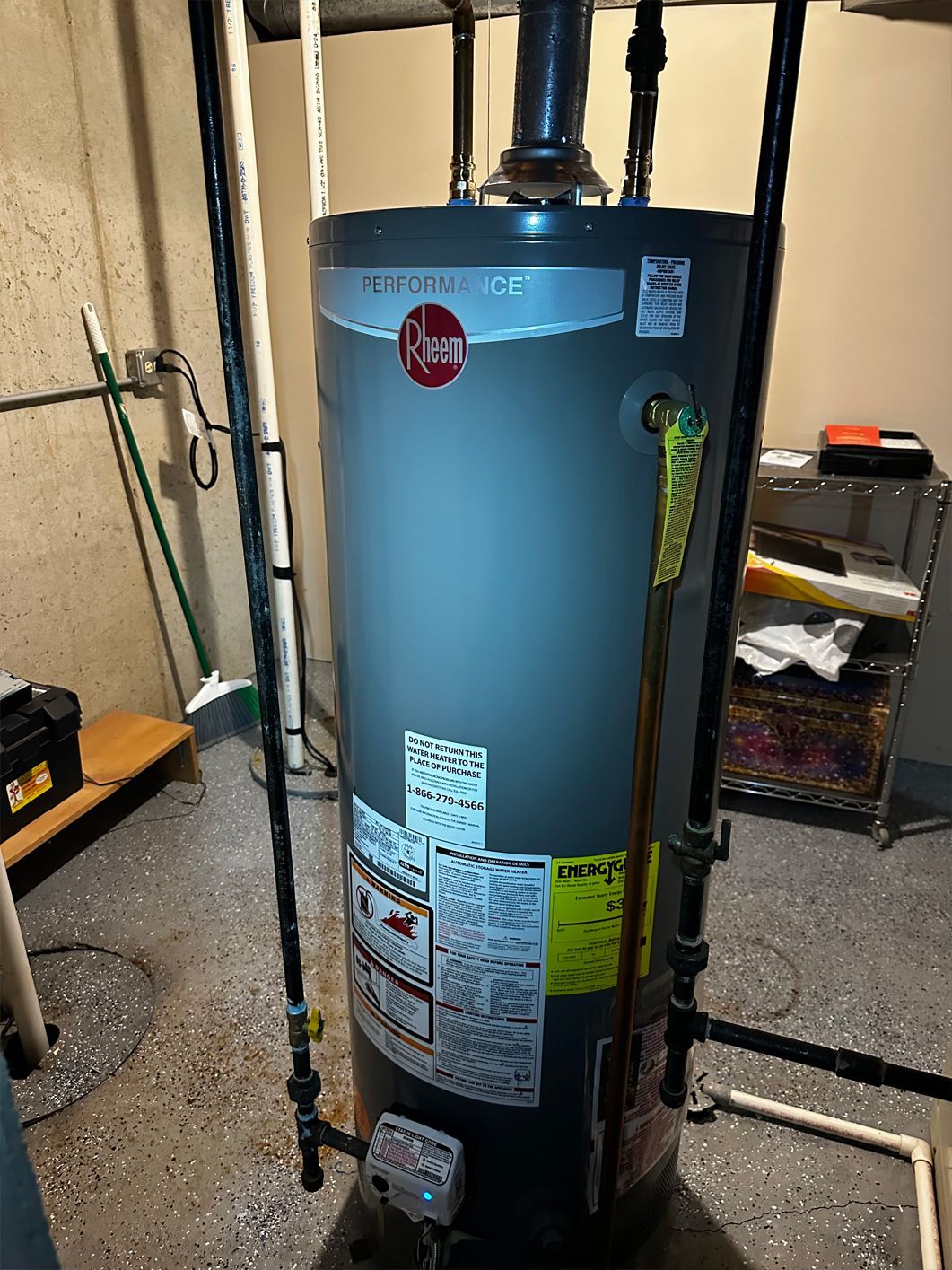 Rheem water heater in a basement setting. Gray tank with attached pipes and labels. Concrete floor.