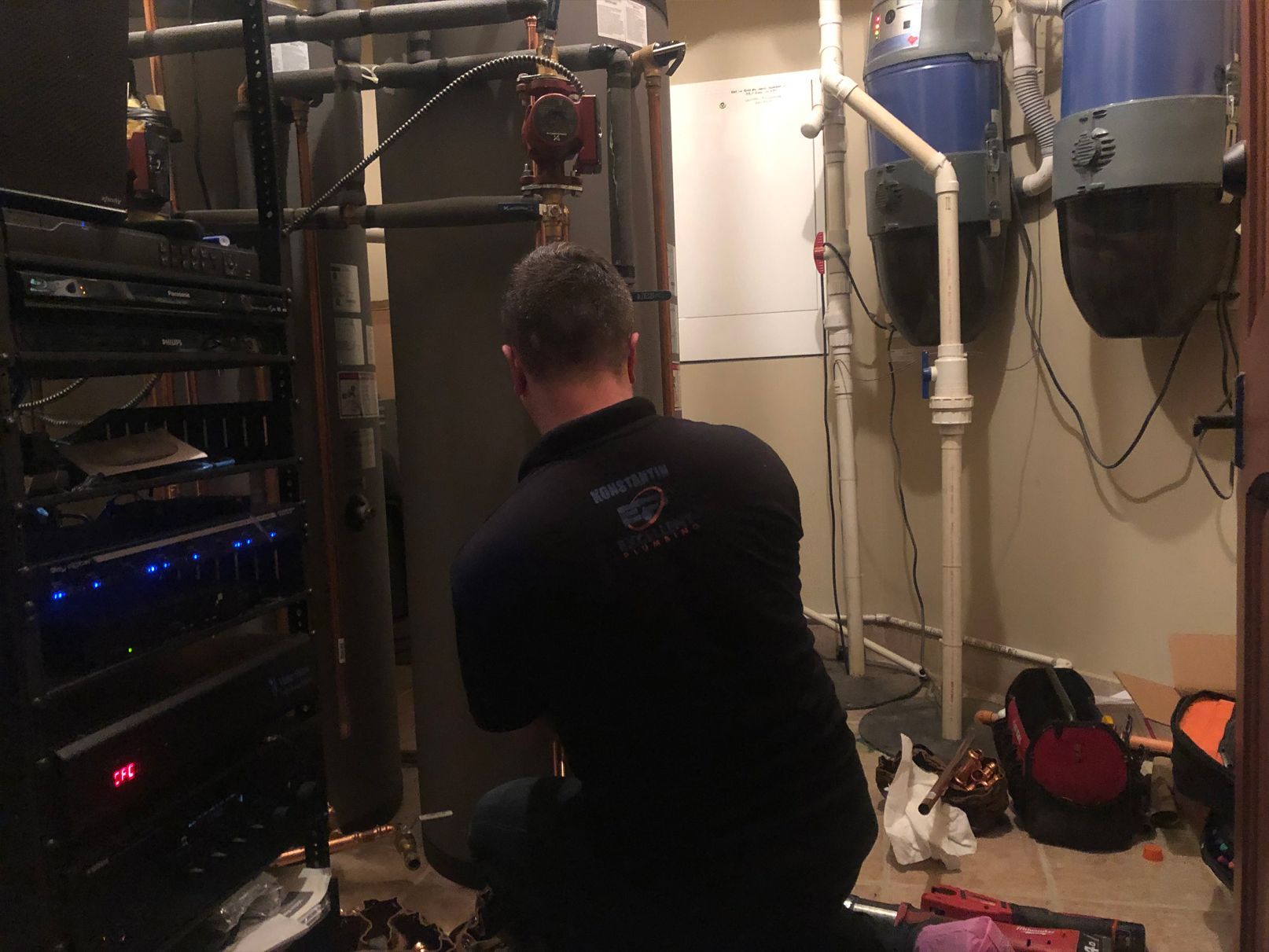 A person in a black shirt works on plumbing in a utility room. Other equipment is also visible.