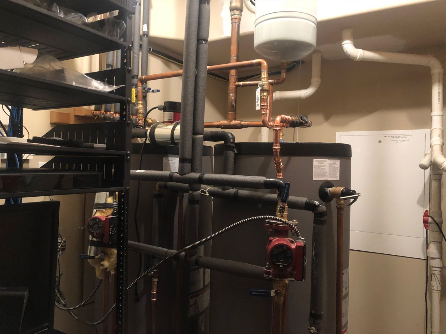 Plumbing system with copper pipes, a water tank, and red pumps, indoors, near a metal shelf.