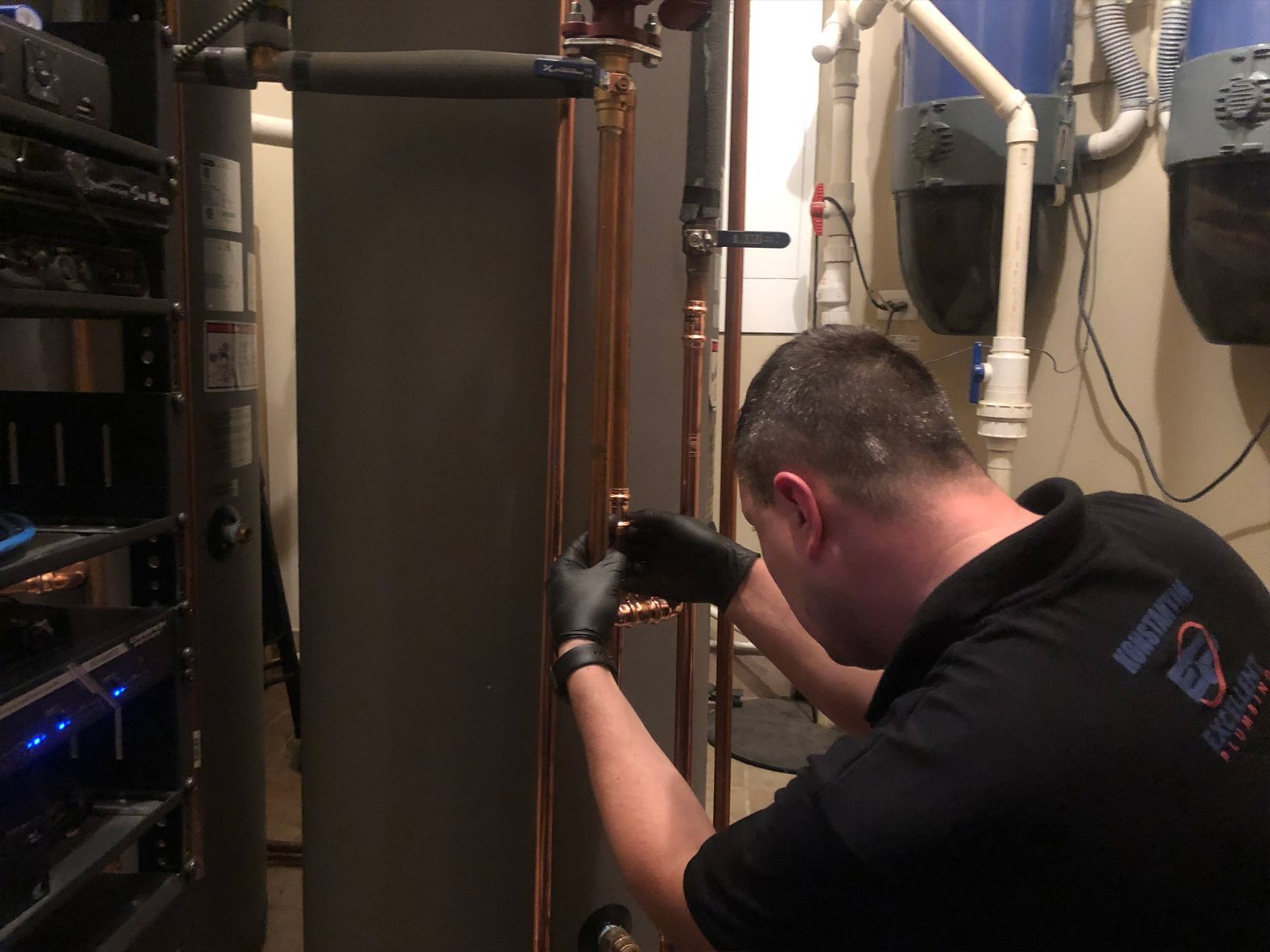 Plumber in black gloves, inspecting copper pipes near a water heater. Interior setting.