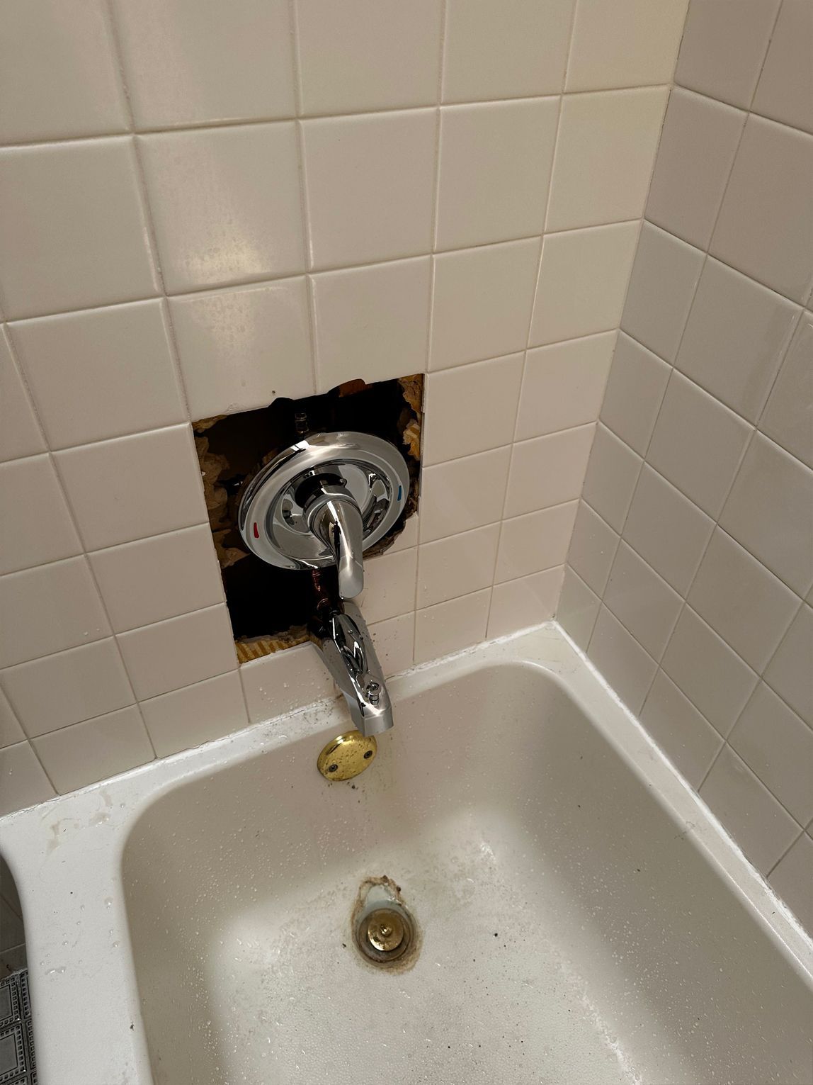 A bathtub faucet and handle are set in a tiled wall with a missing tile section. The tub is white.