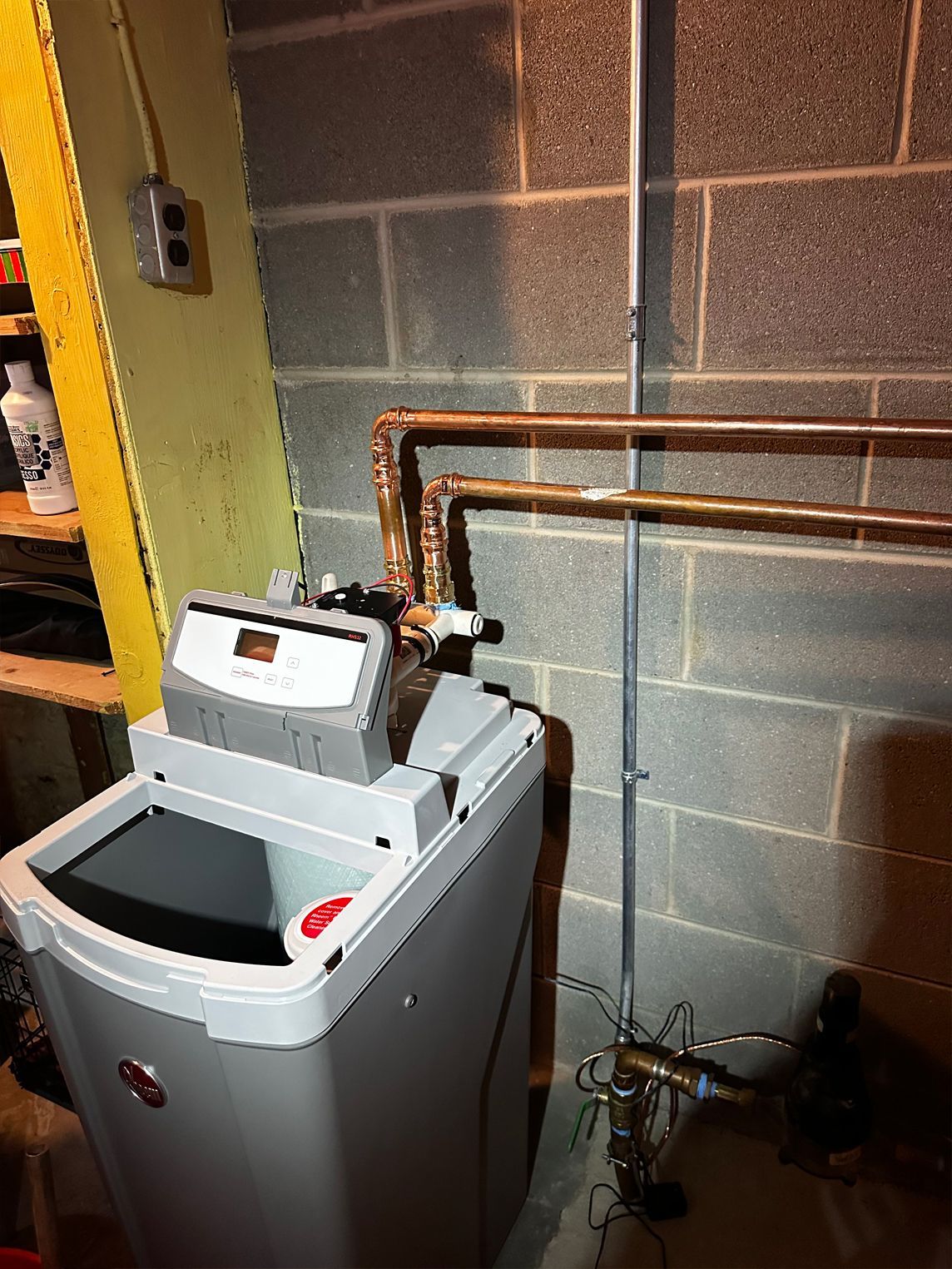 Water softener unit with copper pipes in a basement against a concrete block wall.