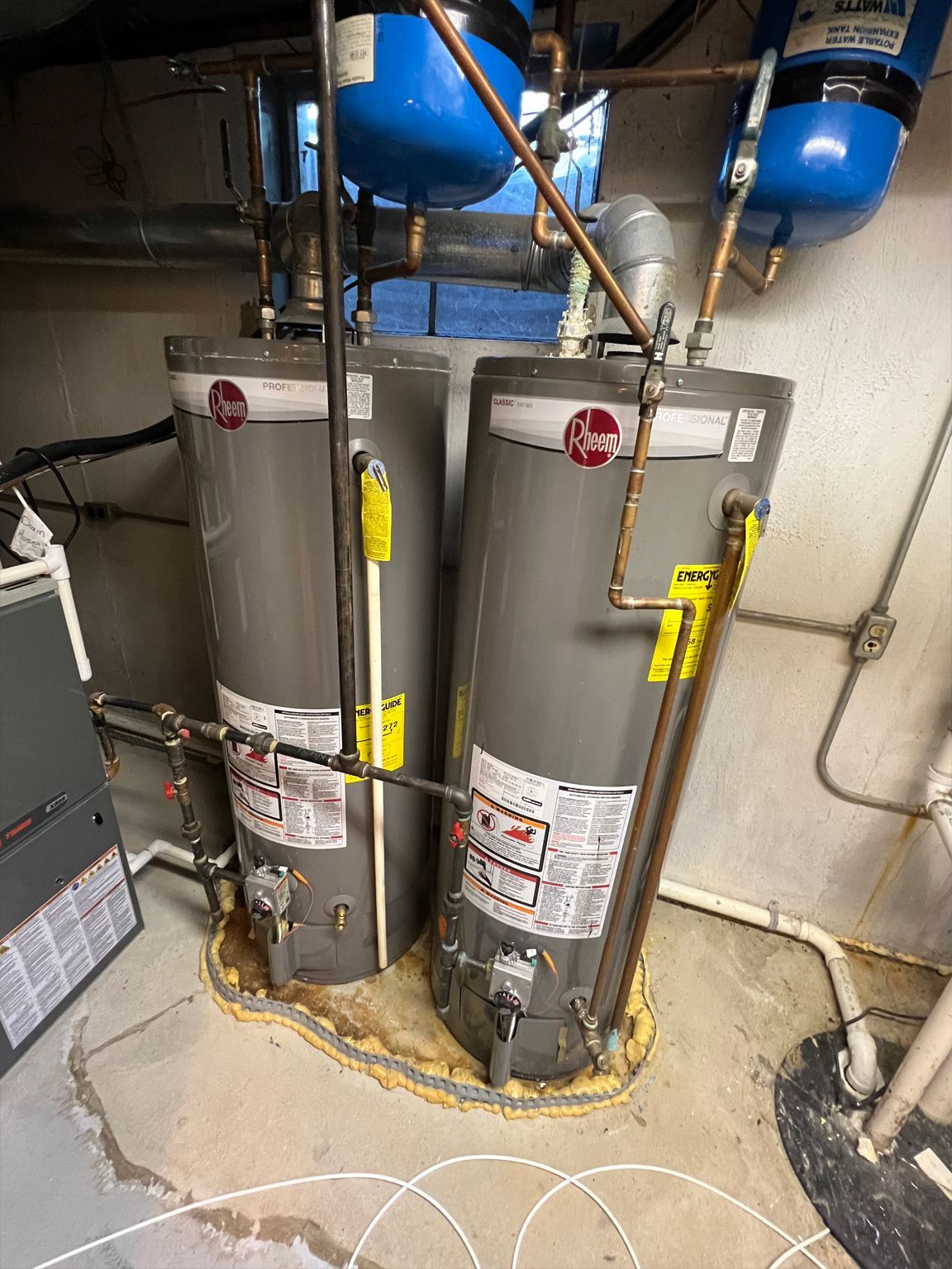 Two gray Rheem water heaters in a basement, with copper pipes and blue pressure tanks above.