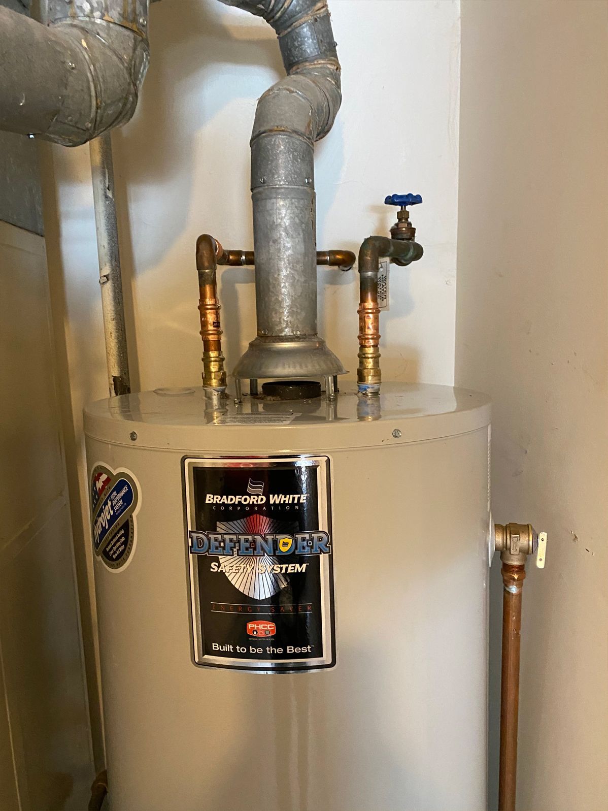 A gray water heater with black labeling and copper pipes, topped with a vent, in a white-walled room.