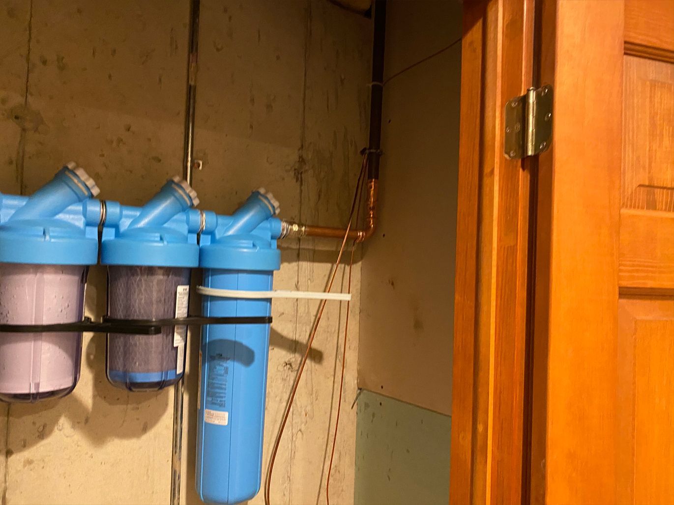 Water filter system on a concrete wall with copper piping, near a wooden door. Blue filters and metal frame.
