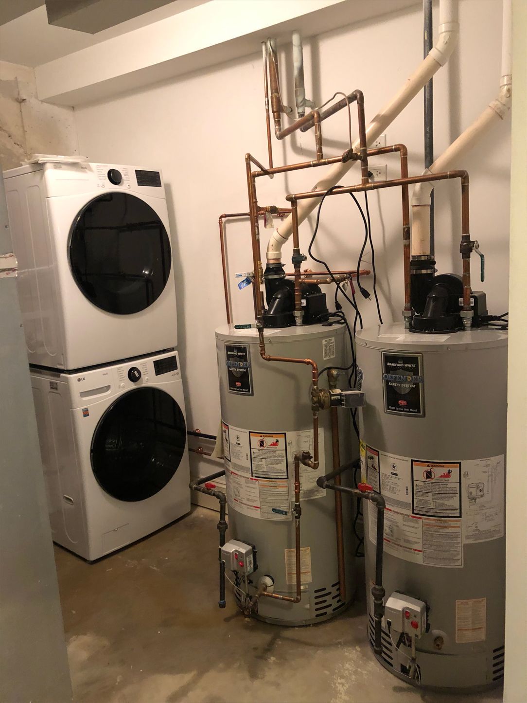 Stacked white washer and dryer next to two tall water heaters with copper pipes in a utility room.