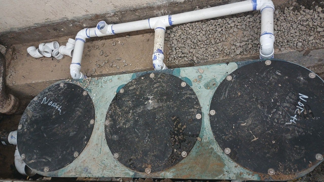 Three circular black lids with white PVC piping, embedded in a concrete trench.