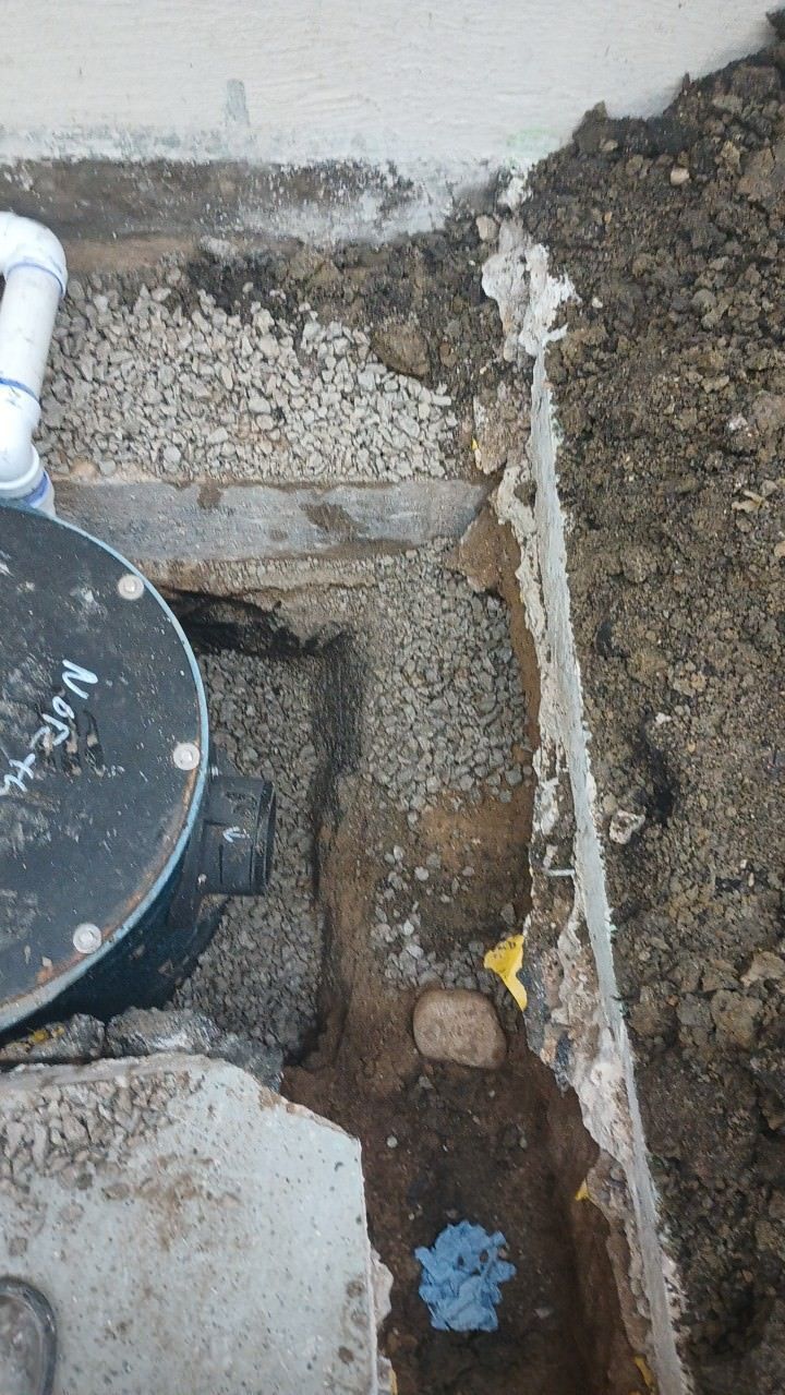 Trench dug along a concrete foundation, revealing pipes, gravel, and fittings; dirt surrounds the excavation.