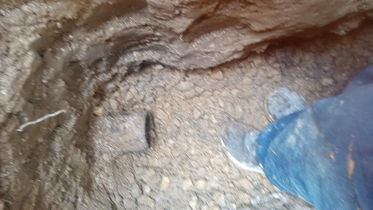 Foot in a boot next to a rectangular metal object embedded in rough, earthy ground.