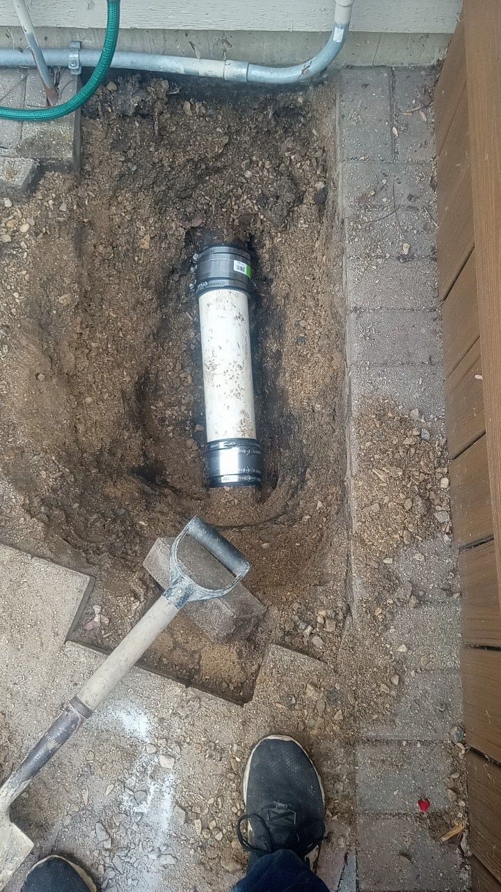 Open hole in dirt with exposed pipe, shovel nearby, next to a concrete surface.