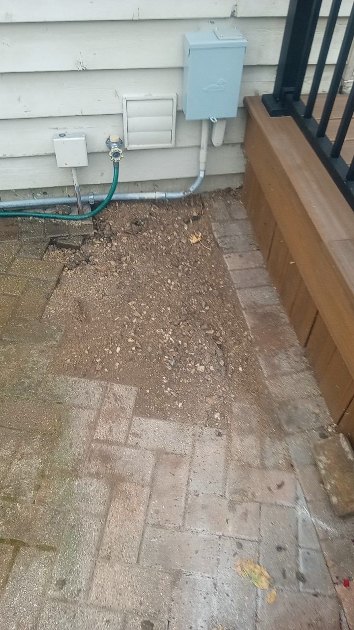 A patio area with missing brick pavers, dirt, and water hose near a building.