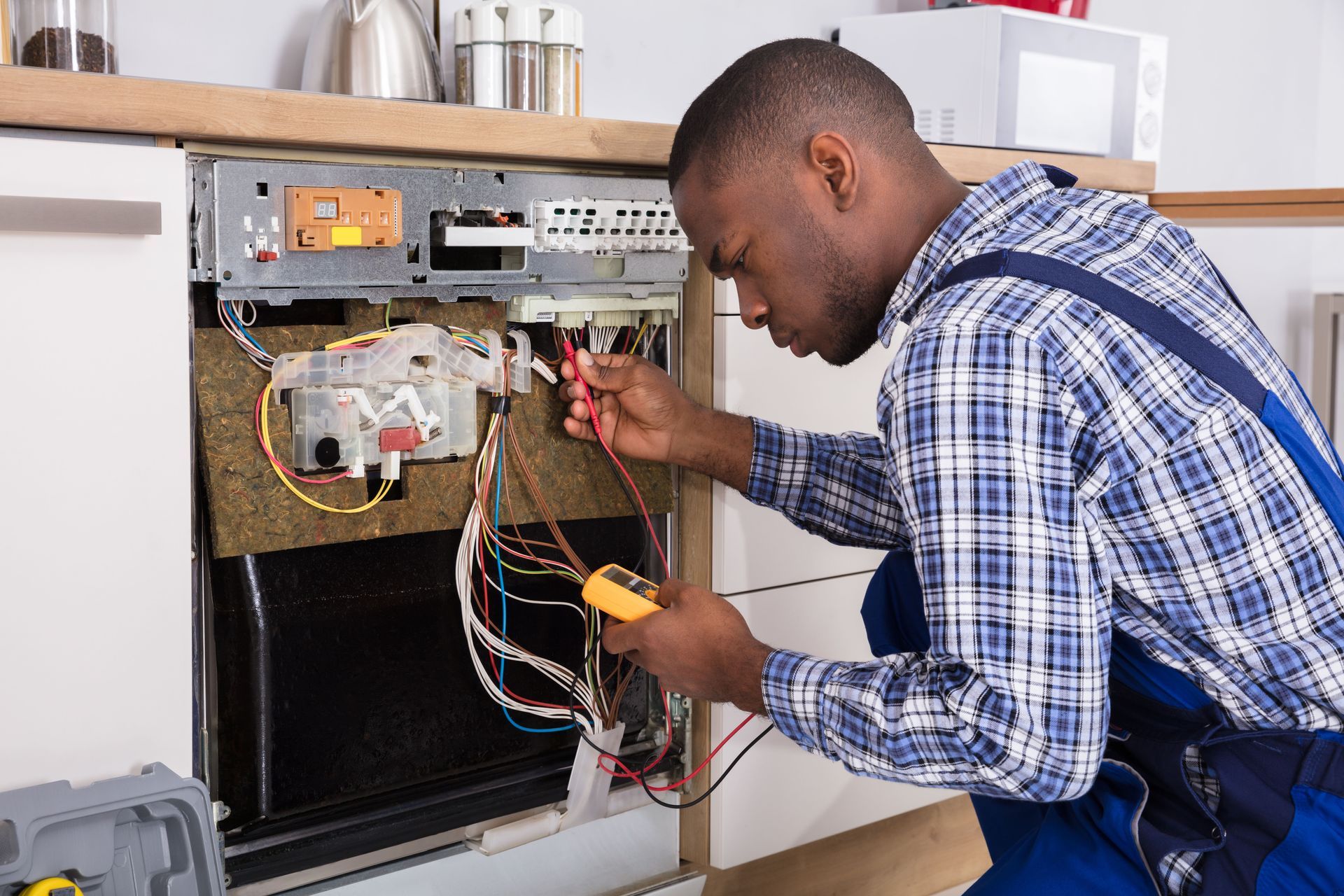 Man in blue coveralls using a multimeter to inspect wiring inside a dishwasher.
