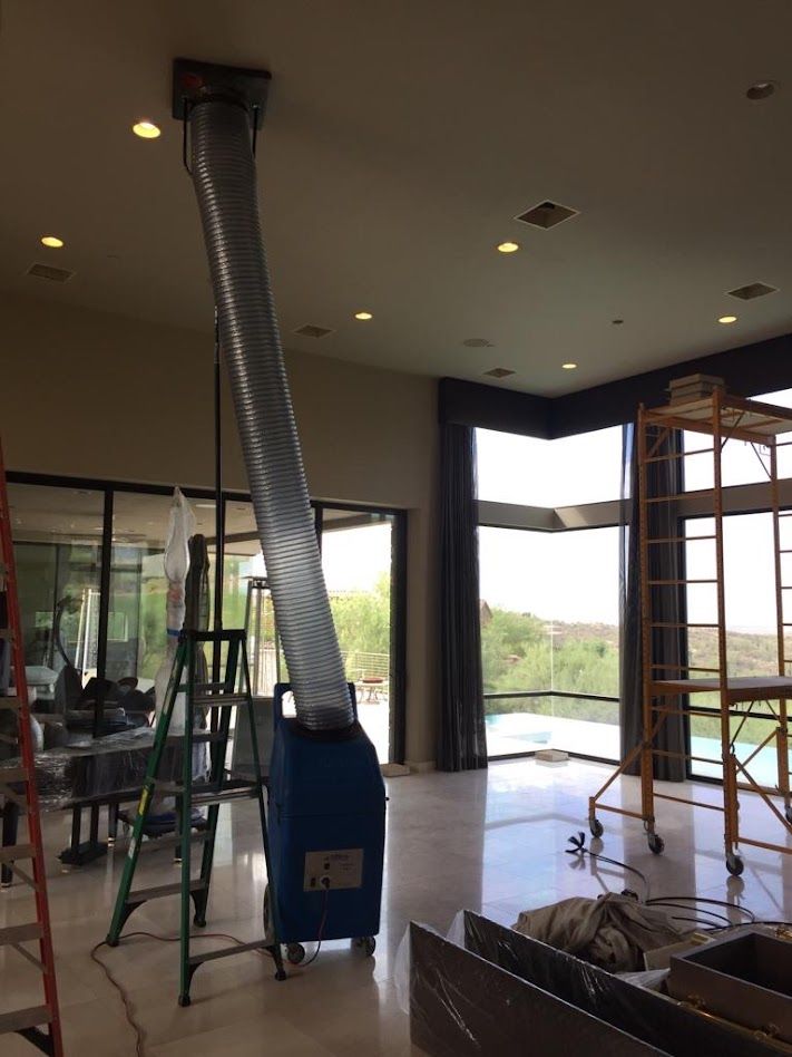 Blue dehumidifier with a silver hose extending to the ceiling in a room with large windows and construction equipment.