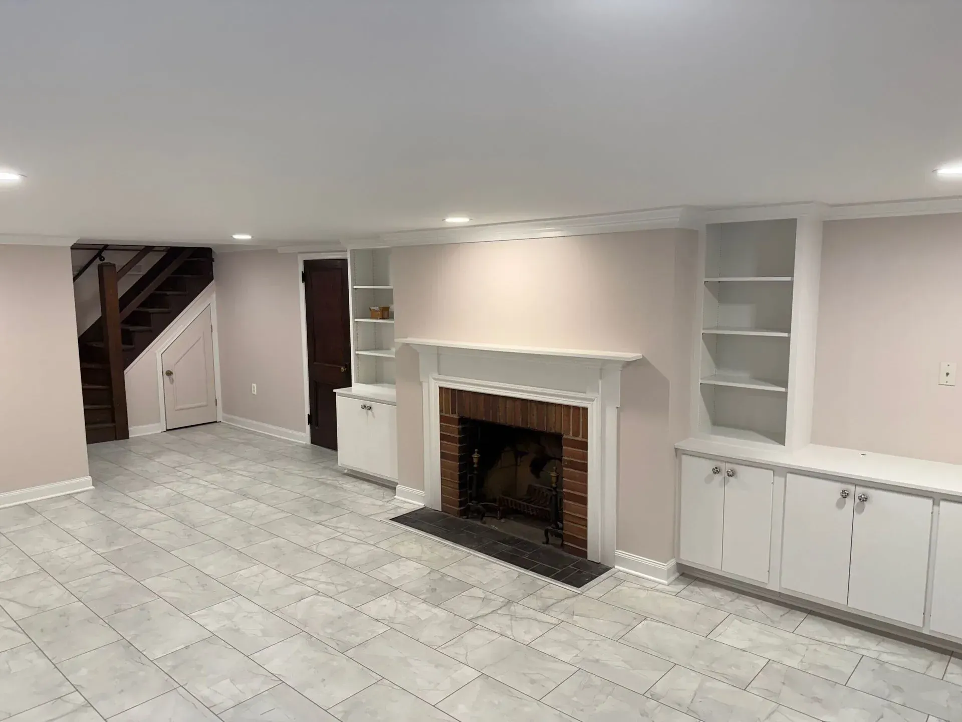 Empty basement with fireplace, built-in shelves, stairs, and tiled floor; pink walls and white trim.