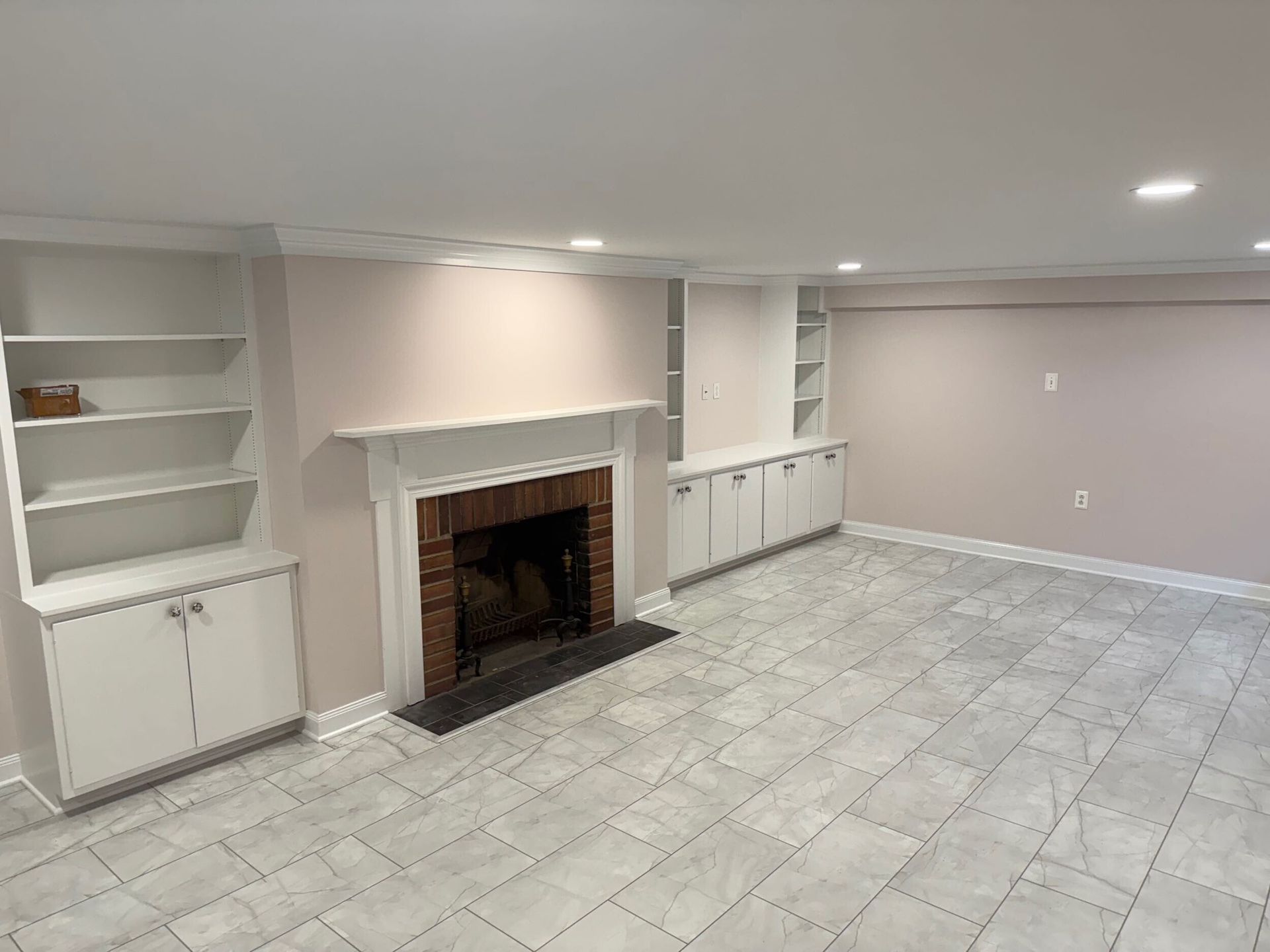 Empty basement room with fireplace, built-in white cabinets, shelves, pink walls, and gray tile floor.