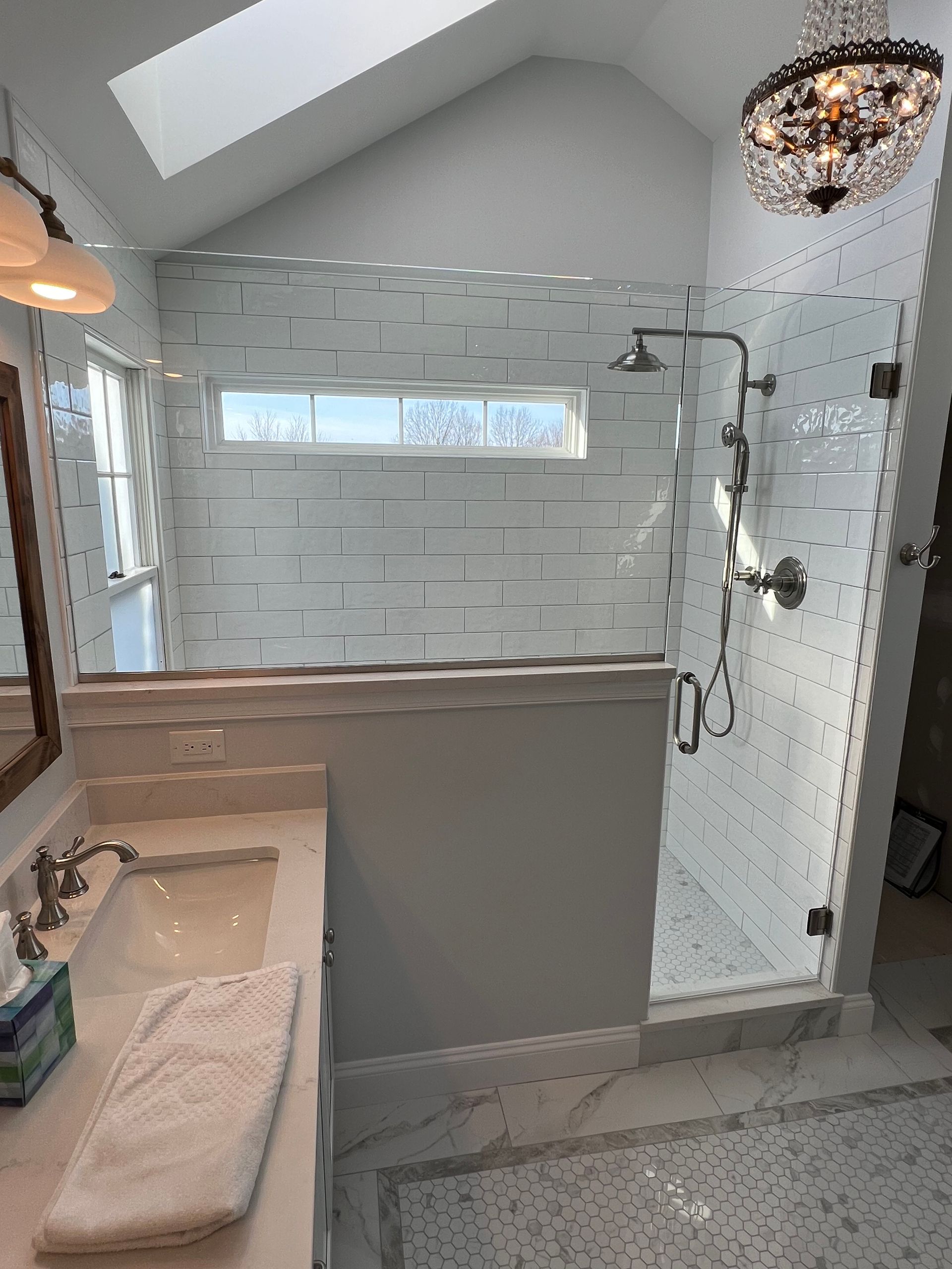 A bathroom with a shower , sink , mirror and chandelier.