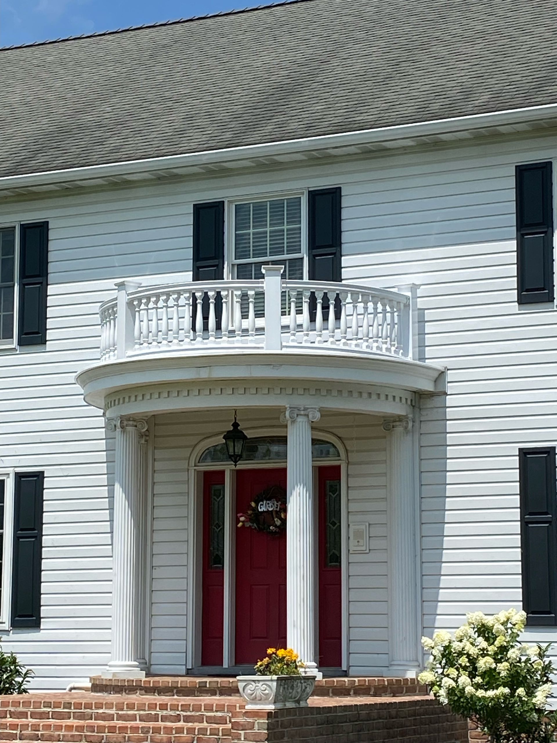White house with a red door, small balcony, and black shutters. Sunlit exterior with stone steps and flower pot.