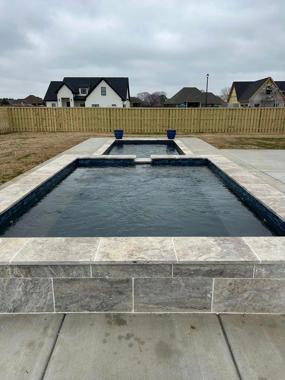 A rectangular pool with a raised edge, surrounded by concrete. Backyard setting with homes and a fence in the background.