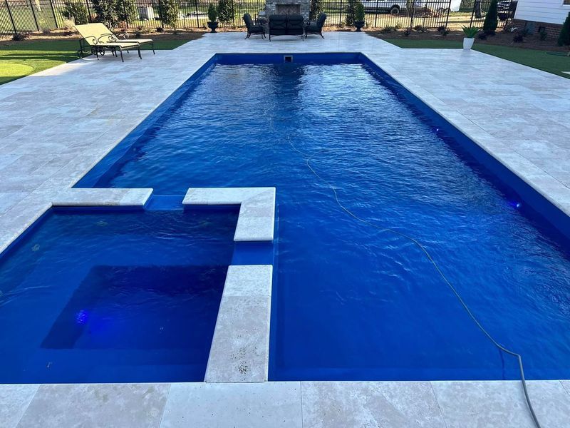 Rectangular pool with attached spa, surrounded by light-colored stone patio. Clear blue water reflects the sky.