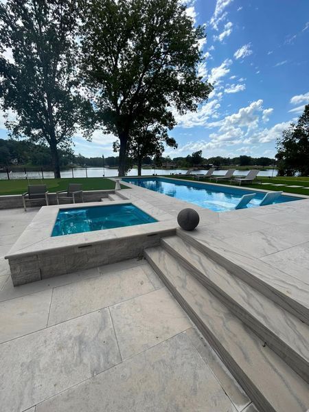 Outdoor pool with rectangular spa, stone patio, and distant lake under a blue sky.