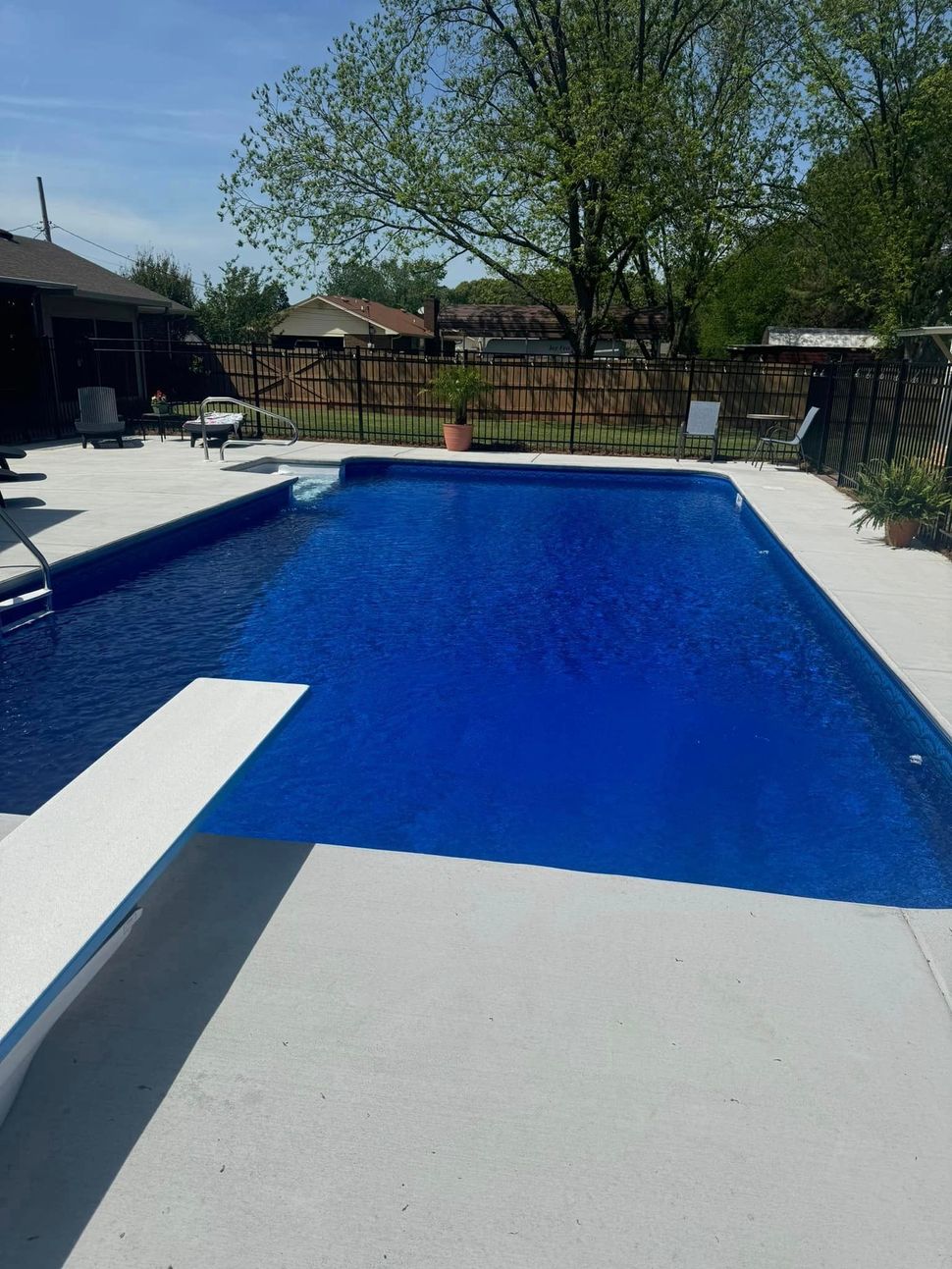 Blue rectangular swimming pool with diving board, surrounded by concrete and a fence in a sunny outdoor setting.
