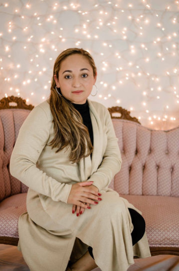 A person with long hair sitting on a pink tufted couch in front of a wall decorated with warm, glowing fairy lights.