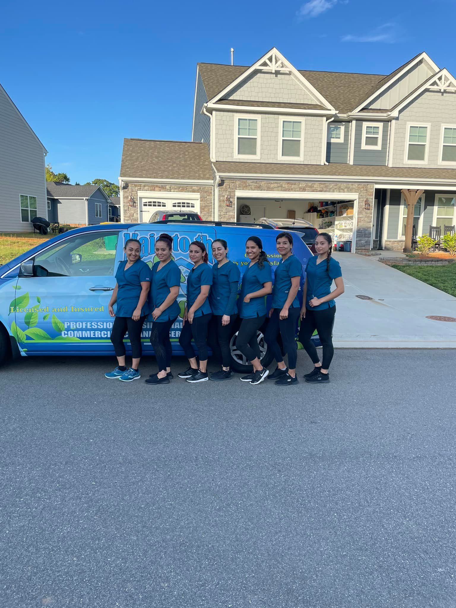 Seven people in matching teal uniforms stand in a line smiling in front of a branded service van parked near a house.